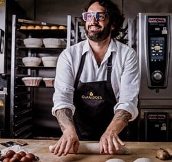A smiling baker wearing glasses and a Claridge's apron is rolling dough on a wooden surface in a professional kitchen, with racks of bread in the background.