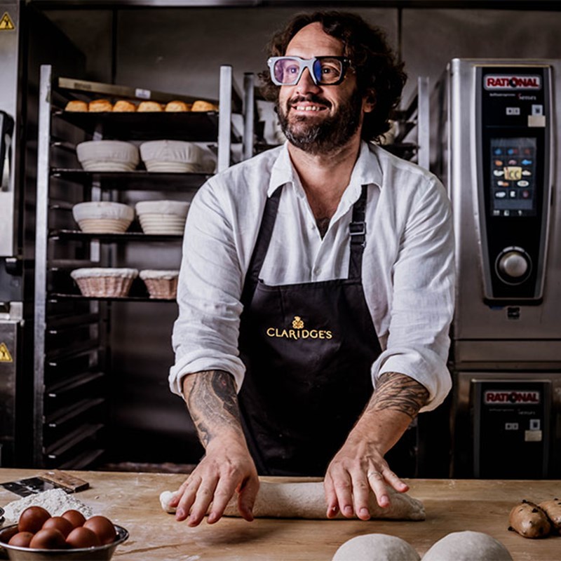 A smiling baker wearing glasses and a Claridge's apron is rolling dough on a wooden surface in a professional kitchen, with racks of bread in the background.
