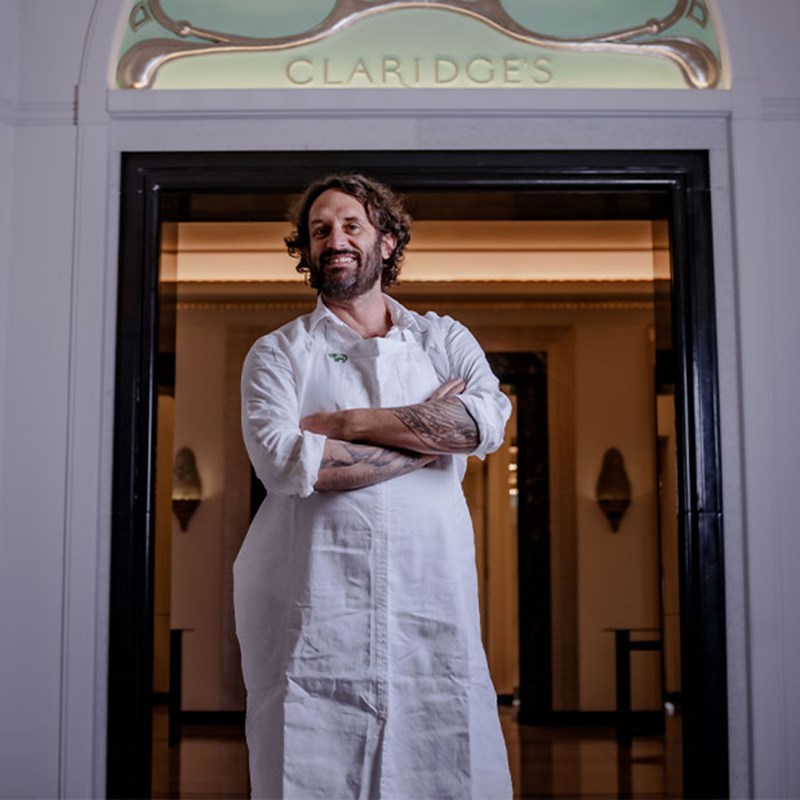 A cheerful baker stands confidently with arms crossed, dressed in a white apron under the elegant Claridge's entrance arch.