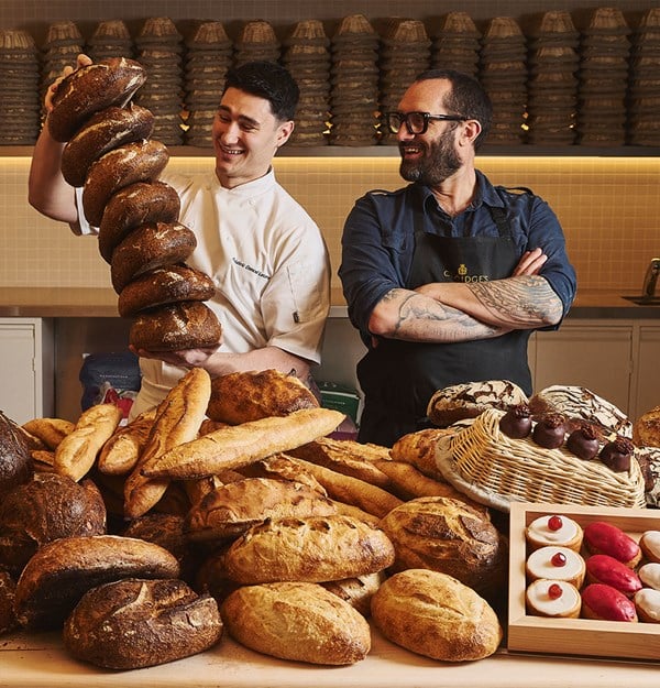 Two bakers smile behind a table of artisanal breads, one holding a tall stack of dark loaves in a warm bakery setting.