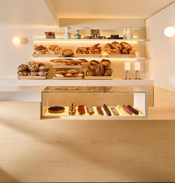 Minimalist bakery interior with shelves of freshly baked breads and pastries above a glass display of cakes.