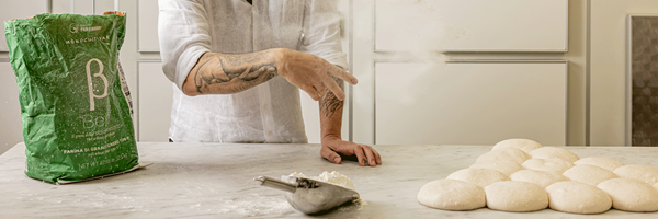 Man flicking flour across a counter beside dough balls, with a flour bag and scoop in view.