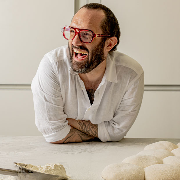 Man in red glasses laughing as he leans on a floured counter beside rounded dough pieces.