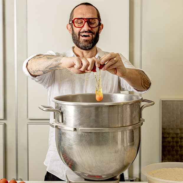 Man cracking an egg into a large metal pot, smiling as he cooks in a bright kitchen.