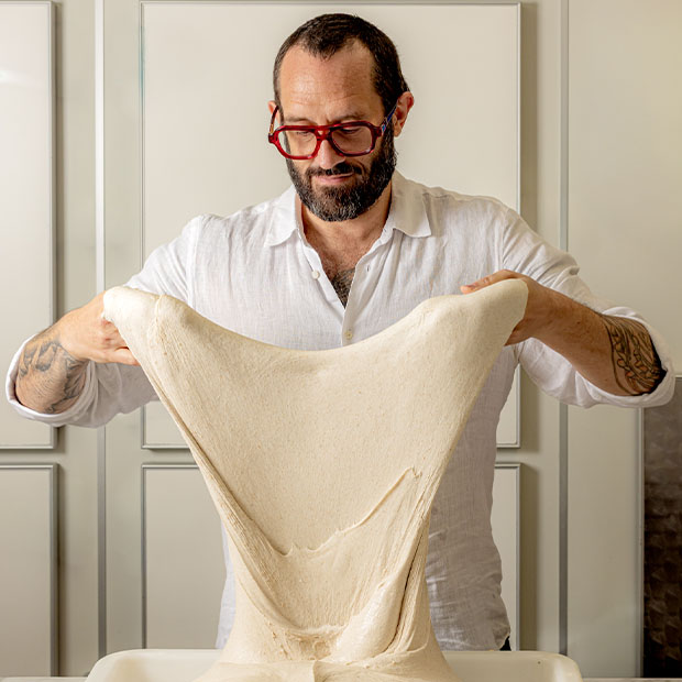 Man stretching a large piece of dough upward between both hands in a kitchen setting.