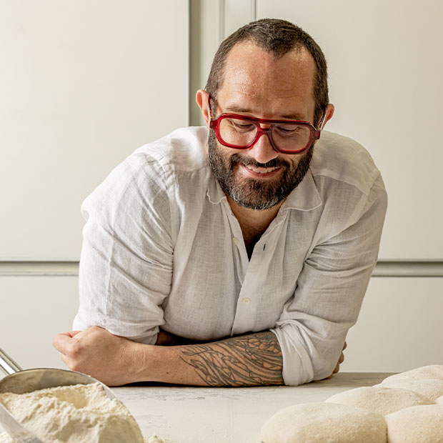 Man in red glasses leaning on a counter, looking at flour and dough with a relaxed smile.
