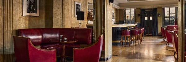 Wide-angle view of Claridge’s Bar featuring glossy red leather banquettes, polished wood floors, and a curved marble bar with red leather stools, framed by gold-toned columns and black-and-white portraits.