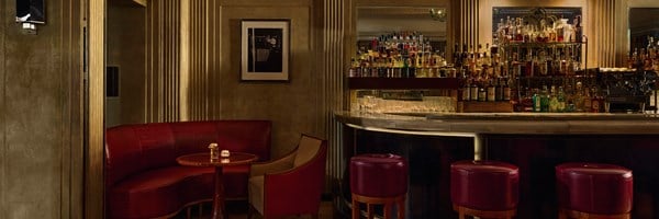 Evening view of Claridge’s Bar showing a softly lit curved bar stocked with spirits, a red leather banquette and armchair in the foreground, and gold-accented walls with art deco details.