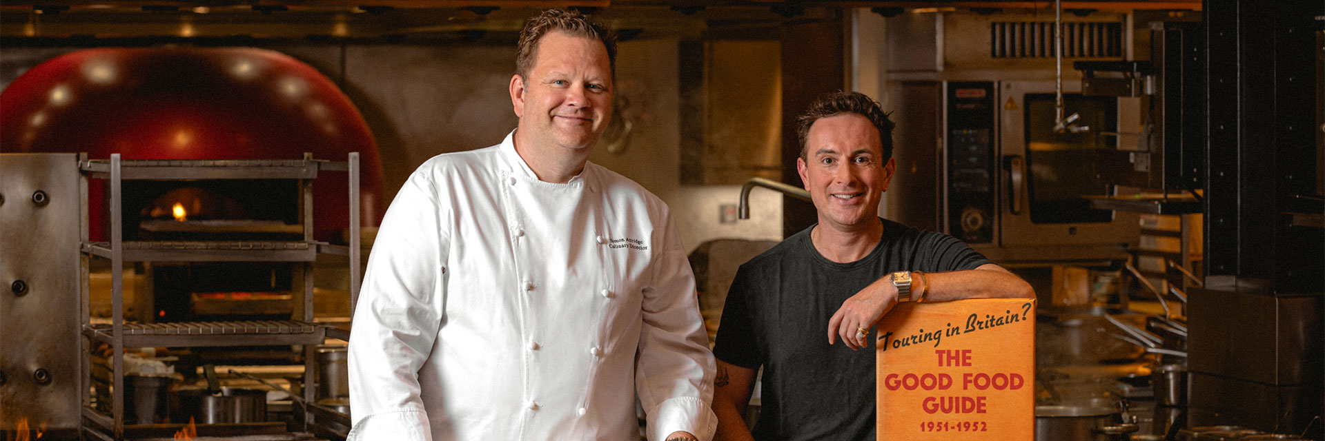 Two men stand in a professional kitchen—one in a chef's coat, the other in a black T-shirt—smiling beside a vintage copy of The Good Food Guide.