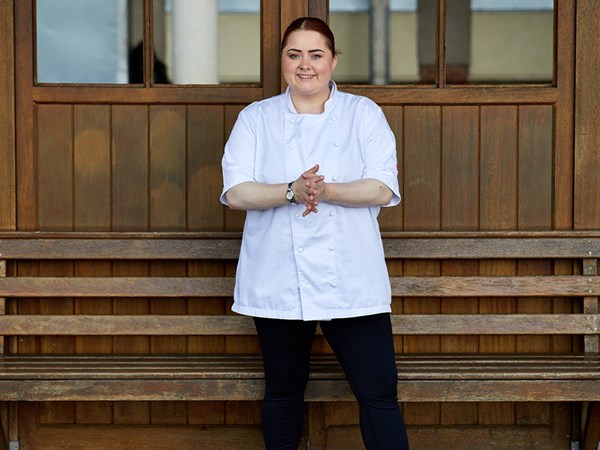 A smiling chef in a white double-breasted jacket and dark trousers stands in front of a wooden bench and panelled wall.