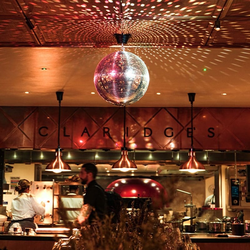 A stylish open kitchen at Claridge’s, illuminated by a dazzling disco ball reflecting light across the space, with chefs at work beneath warm copper pendant lighting.