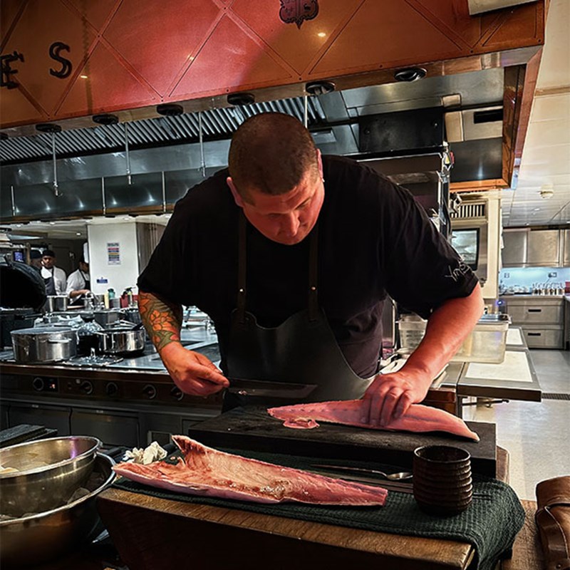 A skilled chef, clad in black with tattooed arms, expertly filleting fresh fish on a wooden board in the refined yet bustling kitchen at Claridge’s.