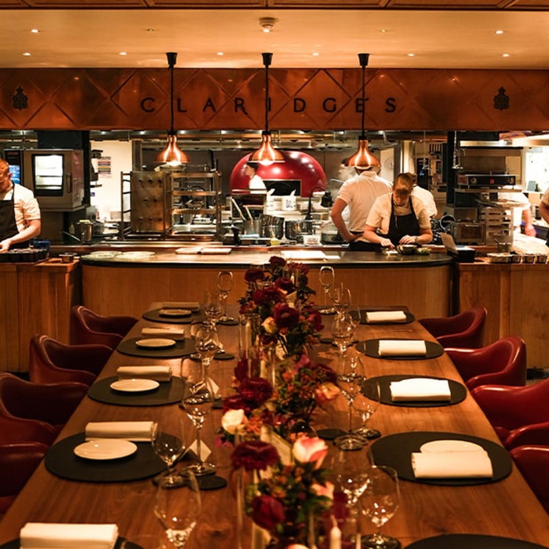An intimate chef’s table at Claridge’s, elegantly set with fine tableware and a floral centrepiece, overlooking the dynamic open kitchen where chefs craft exquisite dishes.