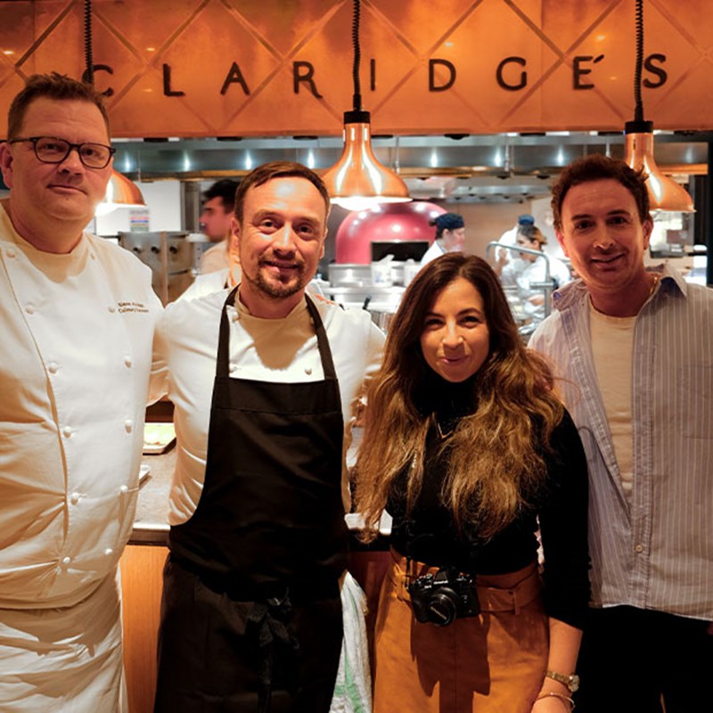A group of chefs and guests, dressed in refined yet relaxed attire, gathered in front of the Claridge’s kitchen, exuding a sense of warmth and culinary camaraderie.
