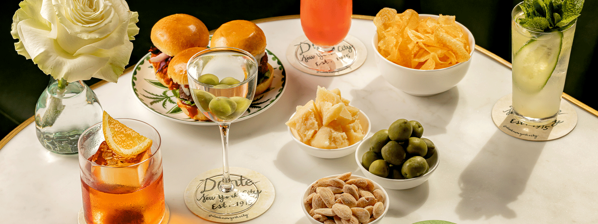 An elegant tabletop spread with gourmet mini burgers, assorted cocktails, and bowls of snacks including olives, almonds, chips, and cheese, accented by a single white rose in a glass vase.