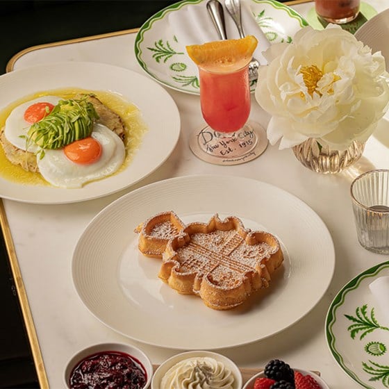 Brunch table setting with a flower-shaped waffle dusted with powdered sugar, a plate of fried eggs with avocado and sauce, a pink cocktail garnished with an orange slice, and small bowls of jam, whipped cream, and fresh berries, on a white marble surface with a white peony in a silver vase.