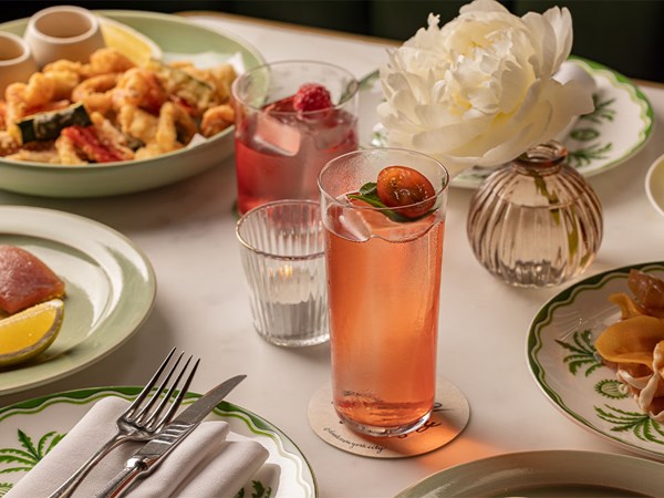A table set with vibrant cocktails, including a tomato and basil-infused spritz, alongside a raspberry-garnished drink, served with a platter of golden fried vegetables, seared tuna, and a blooming white peony in a delicate vase.
