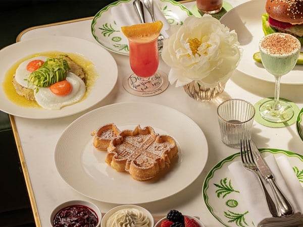 A sunny brunch spread featuring sunny-side-up eggs with avocado, a golden waffle dusted with powdered sugar, fresh berries and whipped cream, a pineapple-topped cocktail, and a green mint dessert drink, arranged with crisp linens and a single white peony.