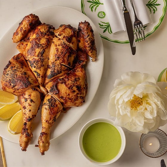 A golden, herb-roasted spatchcock chicken served on a white platter with lemon wedges, accompanied by a vibrant green sauce, floral decor, and an elegantly set place setting.