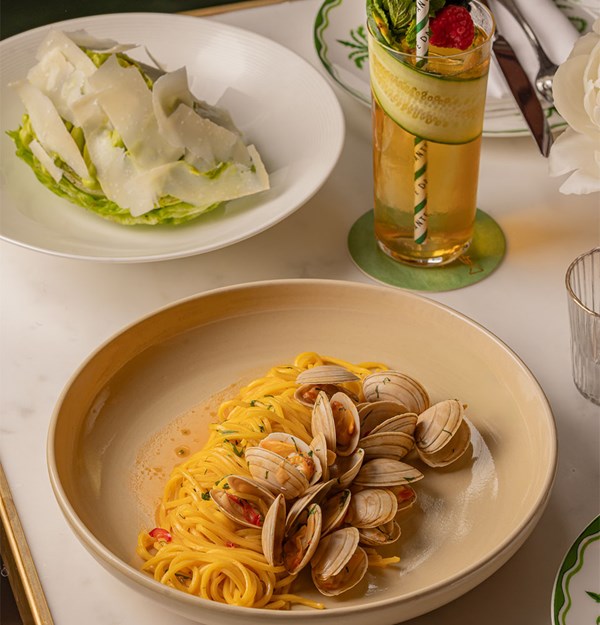 Elegant pasta with clams served in a neutral ceramic bowl, accompanied by a crisp romaine lettuce salad topped with shaved parmesan and a tall cocktail garnished with cucumber and raspberry.