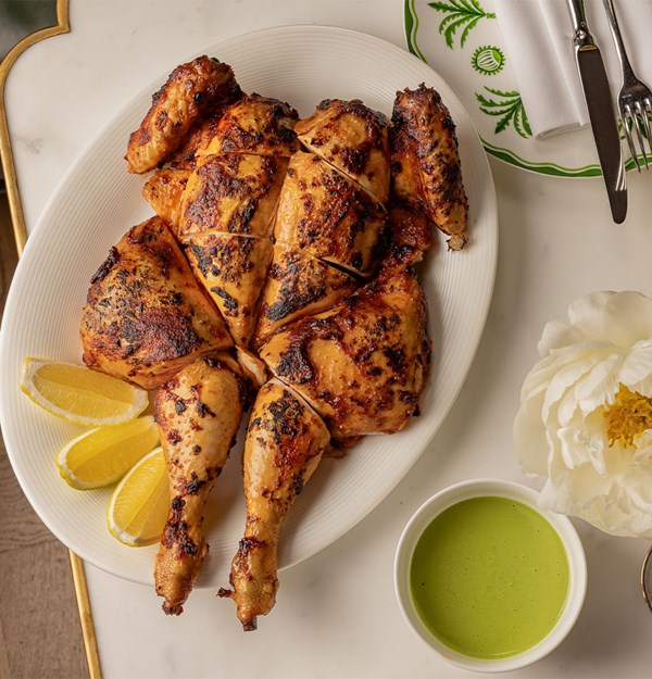 Succulent roasted chicken served whole and expertly carved, paired with lemon wedges and a side of vibrant green sauce, styled with a white peony and fine cutlery.