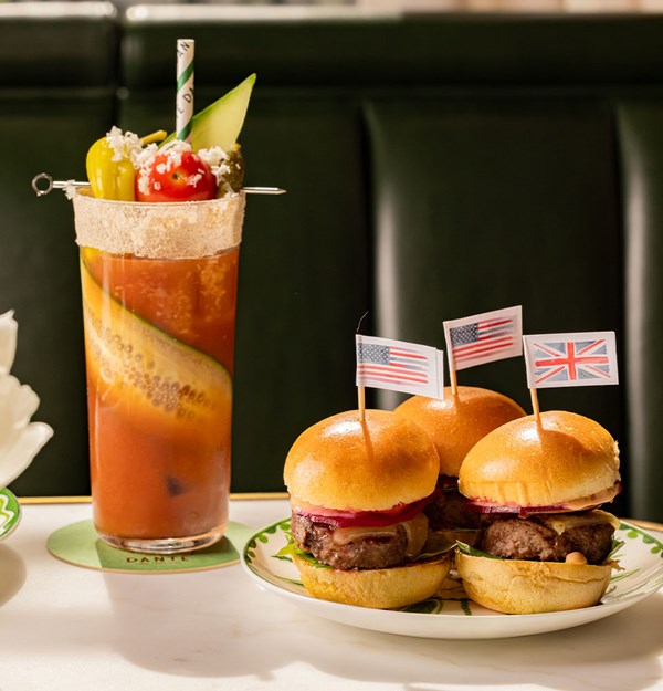 A trio of gourmet mini burgers topped with small American and British flags, accompanied by a tall Bloody Mary cocktail garnished with pickles, cherry tomato, and cucumber ribbon.