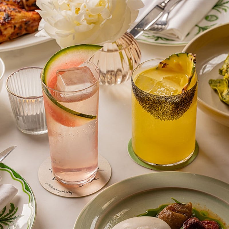 Two vibrant cocktails—one with watermelon and the other with pineapple and a dark rim—set against a table of elegant plates, napkins, and a white peony.