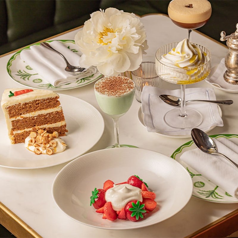 A selection of classic desserts including carrot cake, a pale green dessert cocktail, strawberries with cream, and a swirl of lemon-topped soft serve in a coupe glass.