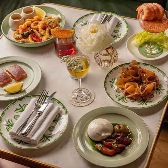 A luxurious table setting featuring Italian antipasti including burrata with roasted vegetables, prosciutto, shrimp cocktail, tuna carpaccio, fried seafood, and two vibrant cocktails, arranged on green-rimmed plates with a white peony in a glass vase.