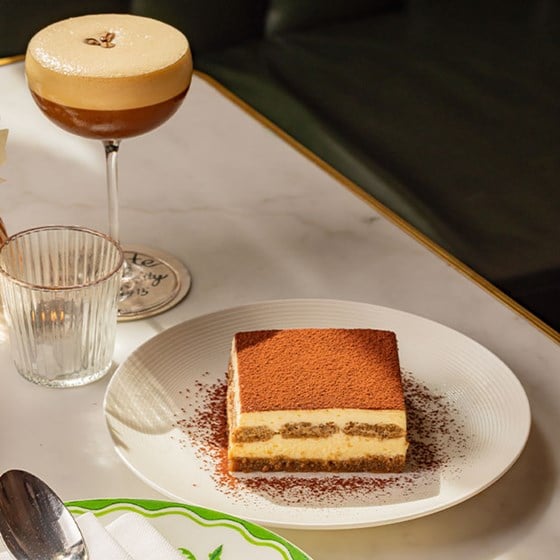 A square serving of tiramisu dusted with cocoa on a white plate, paired with an espresso martini cocktail, resting on a marble table with a lit candle and elegant glassware.