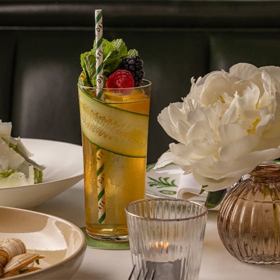 A tall cocktail garnished with a curled cucumber ribbon, mint, and berries, set on a white marble table next to a candle and white peony in a glass vase.