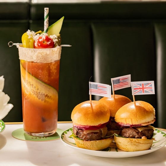 A plate of three gourmet mini burgers topped with miniature flags of the US and UK, served alongside a bold Bloody Mary garnished with olives, tomato, and cucumber.
