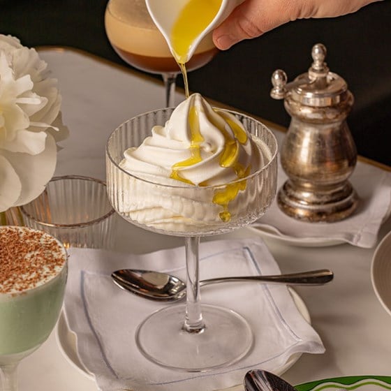 A glass coupe filled with creamy soft-serve gelato being drizzled with golden olive oil, set on a white napkin with a spoon, next to a pepper mill and cocktail glass.