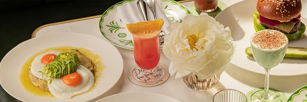 A bright and inviting brunch table featuring sunny-side-up eggs with avocado, a tropical cocktail, a gourmet burger with sesame bun, and a pale green dessert cocktail, styled with a large white flower.