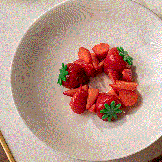 Rotating view of a minimalist dessert made from halved and sliced fresh strawberries arranged in a ring, garnished with green leaf-shaped decorations, presented in a white textured bowl.