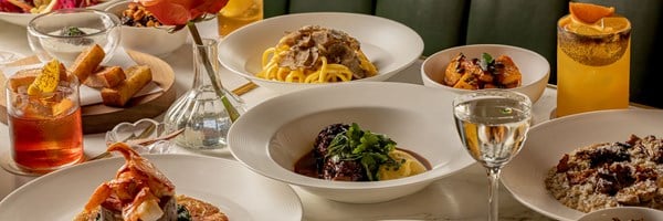 A panoramic view of a table set with Italian dishes including truffle pasta, braised beef, lobster, scallops, risotto, and cocktails, styled with a rose in a vase.