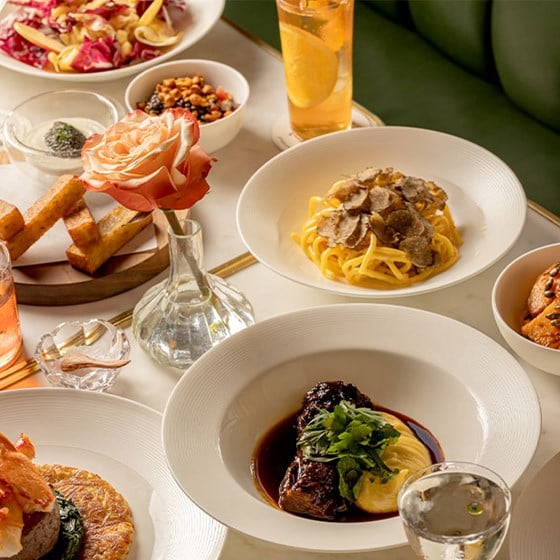 A table filled with Italian dishes, including truffle pasta, braised beef with polenta, salad, breadsticks, and drinks, with a rose in a glass vase at the center.