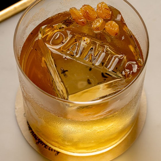 A cocktail in a short glass with a large ice cube engraved with the word “DANTE,” garnished with golden raisins on top.