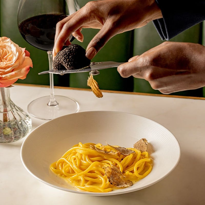 A hand shaves fresh black truffle over a bowl of tagliolini pasta, with a glass of red wine and a rose in a vase nearby.