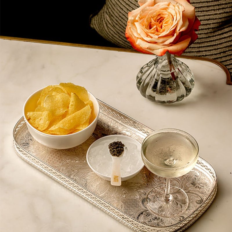 A silver tray with a glass of champagne, a bowl of potato crisps, and a spoonful of caviar on ice, with a rose in a vase placed alongside.