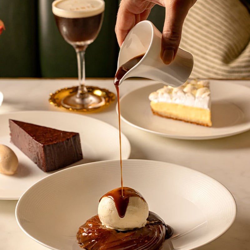 A hand pours warm chocolate sauce over a scoop of vanilla ice cream on a dessert plate, with a slice of chocolate cake, lemon tart, and an espresso martini in the background.
