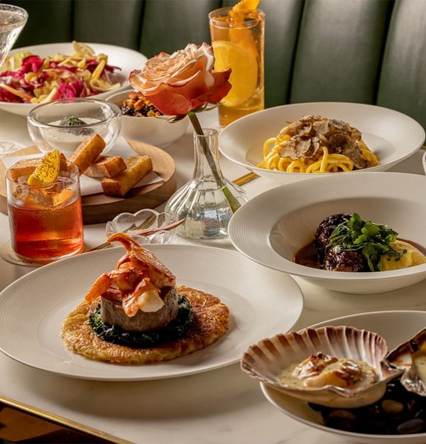 A table spread of fine Italian dishes including truffle pasta, braised beef, lobster with greens, scallops, and cocktails, set around a rose in a glass vase.