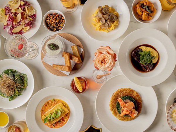 A top-down view of a table filled with Italian dishes, including truffle pasta, braised beef with polenta, lobster with greens, salads, bread, and cocktails, with a rose centerpiece.