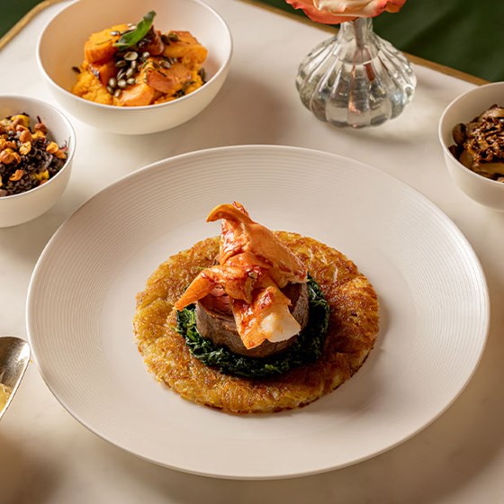 Elegant plated dish of beef topped with lobster on a bed of spinach and crisp potato galette, served with bowls of seasonal sides.