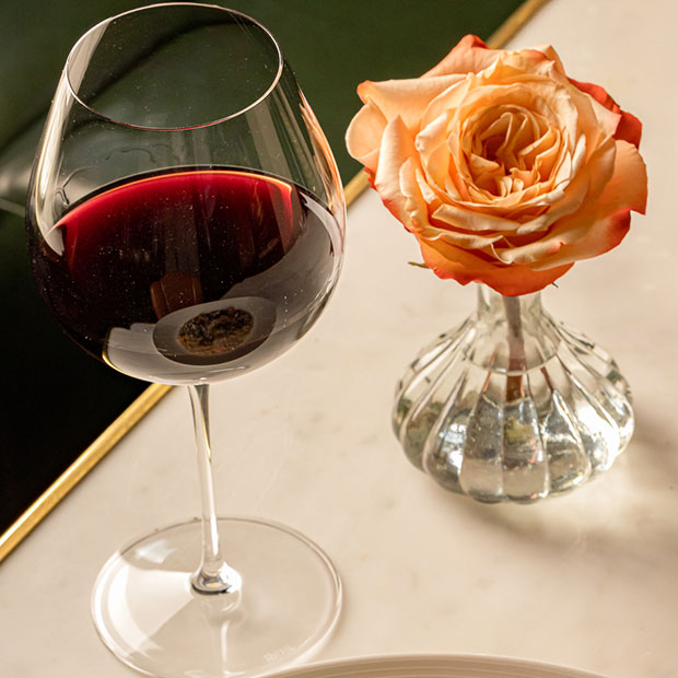 On a marble table sits a glass of red wine on the left and a single orange rose in a bud vase.