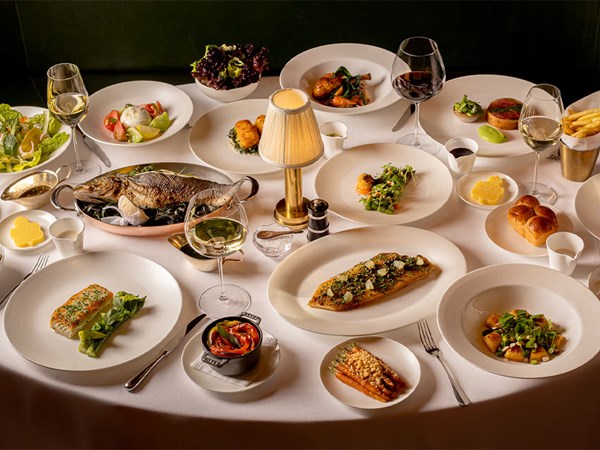 A white tablecloth dining table elegantly set with various gourmet dishes, wine glasses, and a central lamp, showcasing an abundant fine dining Easter spread.