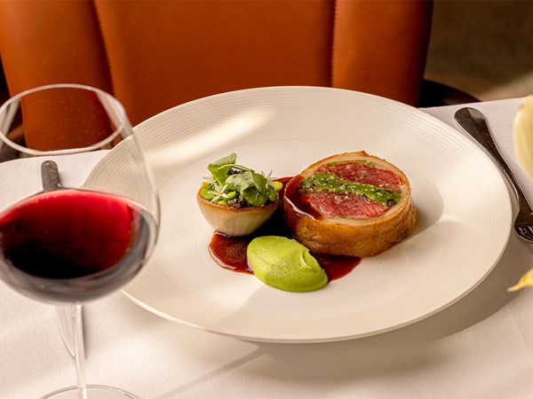 A plated Beef Wellington served with a halved onion garnished with microgreens, pea purée, and a red wine jus, accompanied by a glass of red wine.