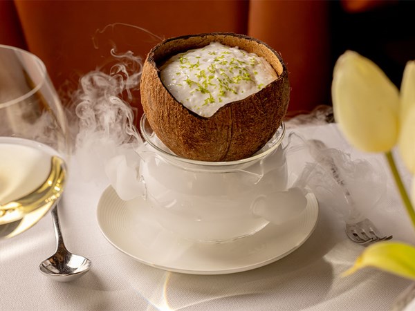 A coconut shell filled with tropical mousse, topped with lime zest and presented over dry ice, with a glass of white wine and tulips beside it.