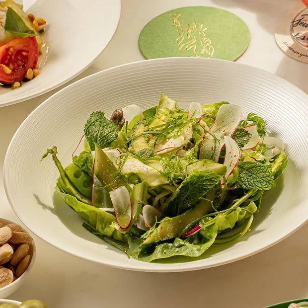 A fresh green salad with shaved vegetables and herbs served in a wide white bowl on a marble table.
