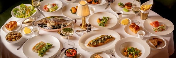 A table set with a variety of gourmet dishes including fish, vegetables, bread, and wine, elegantly arranged on white tablecloth under warm lighting.